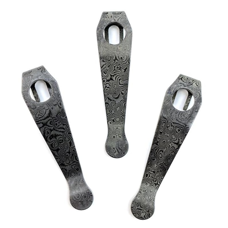 May include: Three metal pocket clips featuring a dark gray and black swirling Damascus pattern. Each clip has a tapered shape with a rounded end and a circular hole at the wider top section, designed for attachment to gear or accessories.