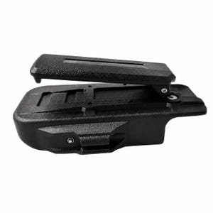 May include: A black, textured plastic holster featuring a rigid, hinged belt clip attached to the top surface. The holster has a molded, contoured shape with visible metal rivets and recessed rectangular sections along the main body structure.
