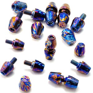 May include: A collection of iridescent metal bolts and caps. The bolts have a threaded end and a hexagonal head. The caps are cone-shaped with a matching iridescent finish of blue, gold, and purple. The items are scattered on a white surface.