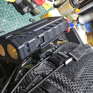 May include: Black tactical device with two yellow lenses, mounted on a black mesh surface with adjustable straps. The device has blue accents and is surrounded by various tools and equipment.