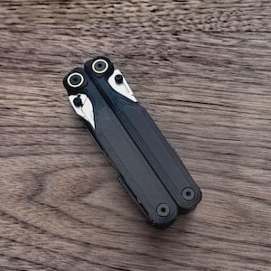 May include: A black and silver multi-tool with a textured handle and silver accents. The tool is closed, showing the handle and the pivot points. The background is a wooden surface.