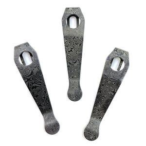 May include: Three metal pocket clips featuring a dark gray and black swirling Damascus pattern. Each clip has a tapered shape with a rounded end and a circular hole at the wider top section, designed for attachment to gear or accessories.