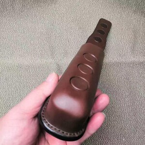 Puede incluir: Un objeto de cuero marr&oacute;n con tres cortes ovalados y una base cosida. El objeto se sostiene en una mano, mostrando su forma y textura. El art&iacute;culo parece ser un asa o agarre.