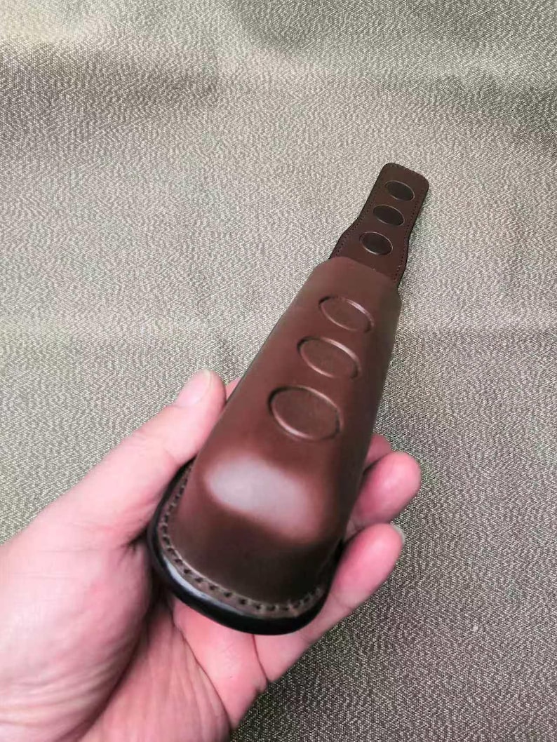 Puede incluir: Un objeto de cuero marr&oacute;n con tres cortes ovalados y una base cosida. El objeto se sostiene en una mano, mostrando su forma y textura. El art&iacute;culo parece ser un asa o agarre.