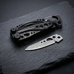 May include: Black Leatherman Skeletool multi-tool and a small folding knife with a patterned blade. The multi-tool is open, revealing its various components. The knife is next to the multi-tool on a dark surface.