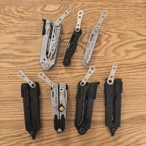 Könnte beinhalten: Eine Auswahl an Multitools in Silber und Schwarz, offen und geschlossen, die ihre verschiedenen Funktionen zeigen. Das Bild ist auf einer Holzoberfläche aufgenommen. Werkzeuge für verschiedene Aufgaben.