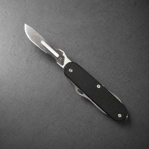 Puede incluir: Un bisturí de acero inoxidable con un mango negro texturizado. La hoja es angular y afilada, con el número "22" grabado en el metal. El mango tiene un pequeño lazo en el extremo.