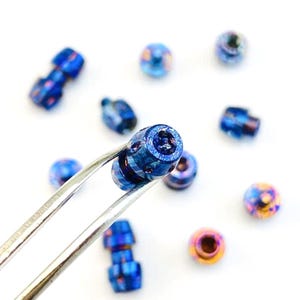 Op de afbeelding: Close-up van verschillende kleine, iriserende blauwe kralen met een metallic glans. Eén kraal wordt vastgehouden door een zilveren pincet, die de cilindrische vorm en meerdere geboorde gaten laat zien. Andere kralen zijn verspreid en tonen een reeks vormen en kleurvariaties.