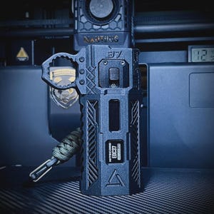 May include: A dark gray, rugged-looking device with the text "NITECORE ED37" and a triangular symbol. It has a handle and a lanyard attached. The device has a digital display and a series of buttons and switches.
