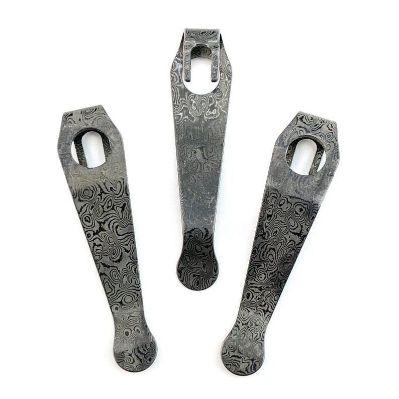 May include: Three metal pocket clips featuring a distinct dark gray and black Damascus steel pattern. Each clip has a tapered body, a rounded tip, and a circular opening at the top end for attachment, showcasing a complex, swirling layered metal design.