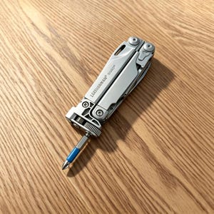 May include: A silver Leatherman Surge multi-tool in a folded position with a Phillips head screwdriver bit inserted. The tool features a metallic finish, visible screws, and a blue-colored ring on the bit, showcasing a compact design.
