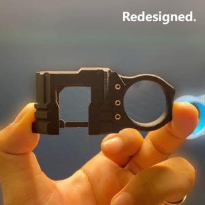 May include: A black, 3D-printed object with a rectangular frame and a circular handle. The object has a geometric design with several small, circular holes and the word "Redesigned." printed at the top.