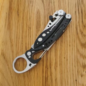May include: A black and silver multi-tool with the brand name "LEATHERMAN" printed on the side. The tool has a carabiner clip and a variety of folding implements. The handle is black with silver accents and a series of circular cutouts.