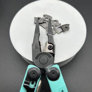 May include: A multi-tool with black pliers and teal handles, featuring the brand name "LEATHERMAN". The tool is open, displaying the pliers, and is accompanied by several small, patterned metal pieces. The background is a light gray circle.