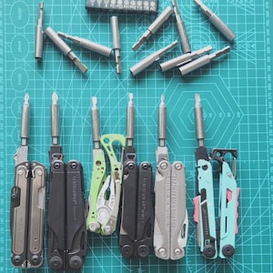 以下が含まれることがあります： 緑色のカッティングマットに配置されたマルチツールとドライバービットのコレクション。黒、シルバー、ティール色のマルチツールがいくつか表示され、それぞれにドライバービットが取り付けられています。さまざまなドライバービットのセットも存在します。