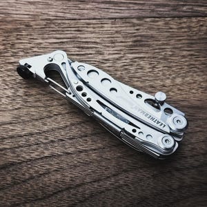 Può includere: Un multiutensile argentato con il marchio "LEATHERMAN" visibile. L'utensile è piegato e si trova su una superficie di legno. L'utensile ha più componenti e una clip.
