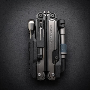 Pode incluir: Uma multiferramenta Leatherman ARC dobrada com acabamento em prata metálica e preto fosco. A ferramenta possui uma chave de catraca visível, um clipe de bolso e um suporte para pontas. O nome Leatherman e o modelo ARC estão gravados no metal.