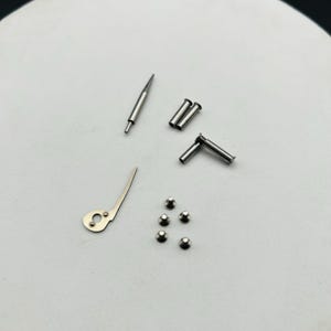 May include: Various small, silver watch repair components, including pins, screws, and a lever, are displayed. The parts are arranged on a light surface, indicating a focus on precision and detail in watchmaking or repair.