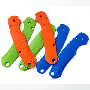 May include: A collection of colorful knife handle scales in orange, green, and blue. Each scale has a textured surface and several holes for attachment. The scales are arranged in a slightly overlapping pattern against a white background.