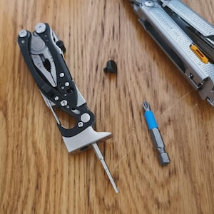 以下が含まれることがあります： 木製の表面に、ペンチ、ドライバービット、黒いハンドルが付いたマルチツールが開いています。青いバンドが付いた別のドライバービットと小さな黒いネジも存在します。ツールはシルバーとブラックです。