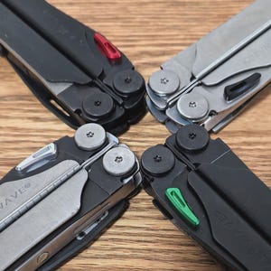 Pode incluir: Quatro multi-ferramentas dispostas em um padrão circular. Duas são pretas com detalhes vermelhos e verdes, e duas prateadas. As ferramentas têm vários componentes, incluindo lâminas. Estão sobre uma superfície de madeira.