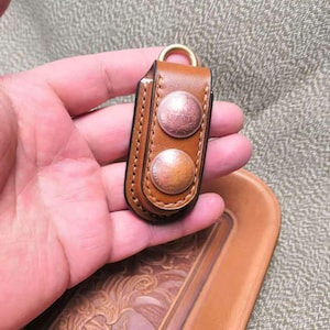 May include: A brown leather key holder with two copper-coloured rivets. The key holder has a gold-coloured ring at the top. The key holder is held in a hand, and a leather tray with a carved design is in the background.