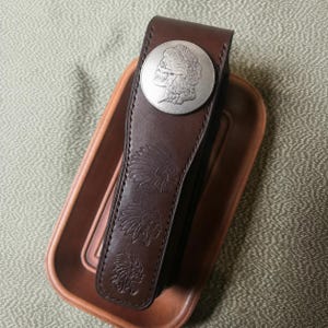 May include: A dark brown leather case featuring a silver-toned circular medallion with a skull profile design at the top. The leather surface is embossed with three repeating images of a Native American headdress in a vertical row along the centre.