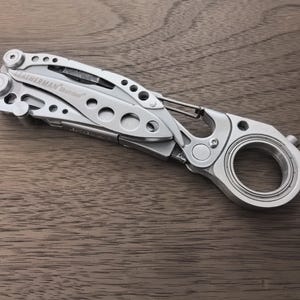 May include: A silver multi-tool with the text "LEATHERMAN Stainless" visible. The tool has multiple components, including a carabiner clip and a large circular opening. It is lying on a wooden surface.