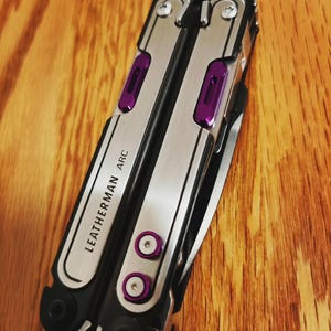 May include: A multi-tool with a silver and black finish, featuring purple accents. The tool has the text "LEATHERMAN ARC" printed on the side. The tool is closed and resting on a wooden surface.
