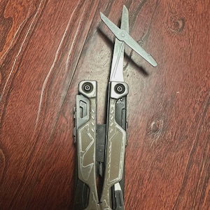 Puede incluir: Una multiherramienta con tijeras abiertas, con una hoja plateada y un mango gris y negro. La herramienta está diseñada con múltiples funciones y se muestra sobre una superficie de madera.