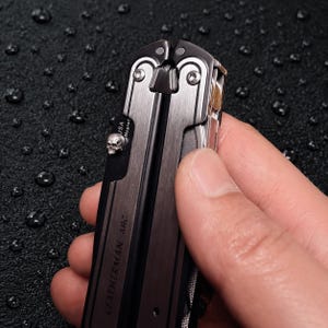 Könnte beinhalten: Ein Leatherman Arc Multifunktionswerkzeug mit gebürstetem, dunklem Metallfinish, einem silbernen, totenkopfförmigen Daumenpin und sichtbarem Text mit der Aufschrift USA und MagnaCut. Das Werkzeug hat ein schlankes, industrielles Design.