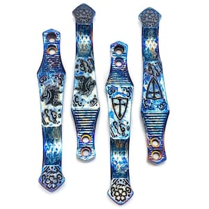 May include: Four decorative metal pieces with intricate designs. The pieces are primarily blue with swirling patterns and accents of other colors. Each piece features a different emblem, including a shield, a horse, and a geometric shape. The ends have floral and geometric patterns.
