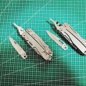 May include: A collection of silver multi-tools and blades are arranged on a green grid surface. The tools include pliers, knives, and other implements, suggesting a focus on utility and versatility. The tools are made of metal.