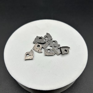 May include: A collection of small, intricately patterned metal pieces, possibly hardware components, arranged on a white, circular surface. The pieces have a swirling, textured design and a dark, metallic finish, suggesting a handcrafted or custom design.