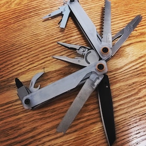 May include: A multi-tool with various implements, including pliers, a saw, a knife, and a file. The tool is gray and silver, with orange accents. It is open, displaying its different functions. The tool is lying on a wooden surface.