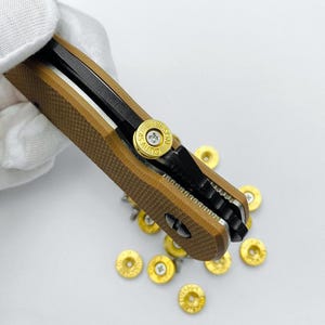 May include: A brown and black folding knife with a textured handle. The knife has a black blade and a gold-coloured accent with the text "EMERSON 45 AUTO". Several gold-coloured bullet casings are scattered around the knife.