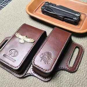 Puede incluir: Estuches de cuero marrón para multiherramientas, uno con un emblema de águila y otro con un emblema de cabeza de indio. Una multiherramienta negra descansa en una bandeja de madera. Las fundas tienen un asa y están cosidas con hilo marrón.