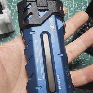 May include: A blue and black flashlight with the text "SK-05 PRO" and "LOOD GEAR". The flashlight has a rectangular design with a black top and a silver accent. The serial number is SN:L 013135.