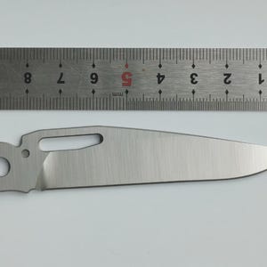 May include: A stainless steel knife blade with a curved edge, a hole, and a rectangular cutout. A metal ruler with millimeter markings is above the blade. The blade is approximately 4 inches long.