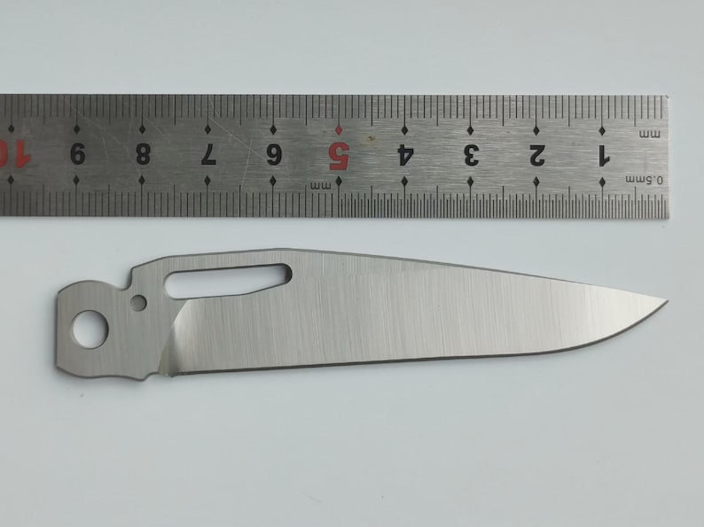 May include: A stainless steel knife blade with a curved edge, a hole, and a rectangular cutout. A metal ruler with millimeter markings is above the blade. The blade is approximately 4 inches long.