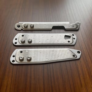 May include: Three silver metal plates with a textured surface and small circular holes. Each plate has several small, round, silver-colored rivets. The plates are arranged on a dark brown wooden surface.