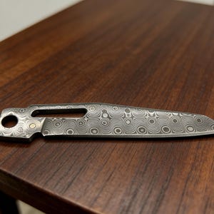 May include: A Damascus steel knife blade with a unique swirling pattern. The blade has a hole near the handle and a small gold-colored accent. The knife is resting on a dark wooden surface.
