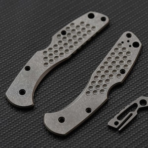 Puede incluir: Dos cachas de metal gris para mango de cuchillo con un patrón texturizado de hendiduras circulares y múltiples orificios para tornillos preperforados. Un pequeño componente de clip de bolsillo de metal gris a juego está colocado al lado.