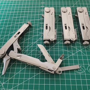 Puede incluir: Una colección de multiherramientas plateadas, algunas abiertas y otras cerradas, dispuestas sobre una esterilla de corte verde. La herramienta abierta muestra alicates, un cuchillo y otros implementos. La marca "Leatherman" es visible.