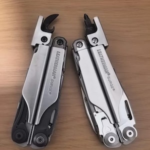 May include: Two silver and black Leatherman Surge multi-tools shown in a folded position. The left tool features an extended strap cutter, while the right tool displays an extended screwdriver bit. Both tools have the brand name engraved on the side.