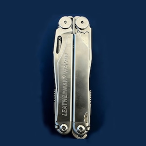 May include: A silver Leatherman Wave multi-tool, shown vertically against a dark blue backdrop. The tool is closed, displaying the "LEATHERMAN WAVE" text. The tool has various edges and tools.