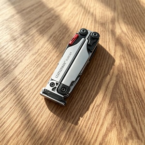 Puede incluir: Una herramienta multifunción Leatherman Surge de color plateado y negro en posición cerrada y plegada. El mango de metal presenta el nombre de la marca Leatherman Surge impreso en negro, con una pequeña pieza roja visible cerca del pivote.