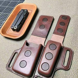 Puede incluir: Una funda de cuero marrón para multiherramientas con tres cierres de botón a presión y una trabilla para el cinturón. Una multiherramienta descansa en una bandeja de cuero a juego. Los artículos se muestran sobre una superficie texturizada.