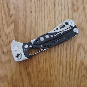 May include: A black and silver multi-tool, with the brand name "LEATHERMAN" visible. The tool is shown against a wooden background. The multi-tool is approximately 10 cm in length and has various functions.