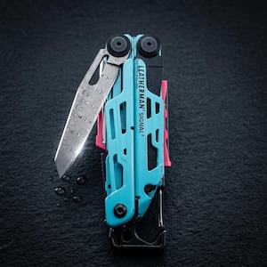 Op de afbeelding: Een Leatherman Signal multitool met een fel turquoise frame met zwarte accenten en een roze detail. Het gereedschap bevat een gedeeltelijk uitgeklapt mes met een opvallend Damascus stalen patroon en de tekst LEATHERMAN SIGNAL op de zijkant.
